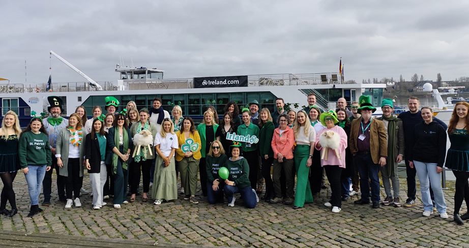 A large group of people dressed in green St. Patrick’s Day–themed clothing posing together on a cobblestone waterfront in front of an Ireland-branded boat.