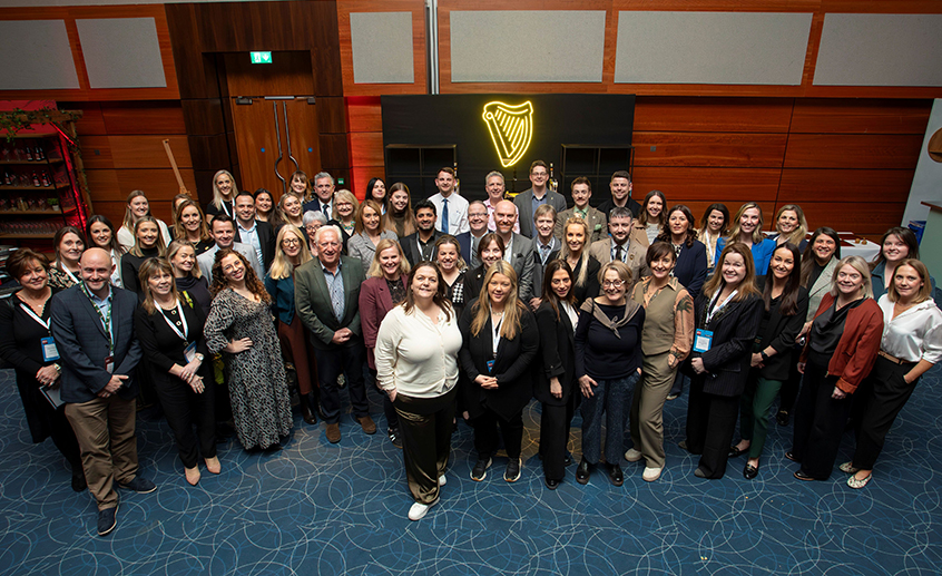 Large group photo of event attendees standing together in a conference room with a lit harp symbol on the wall behind them
