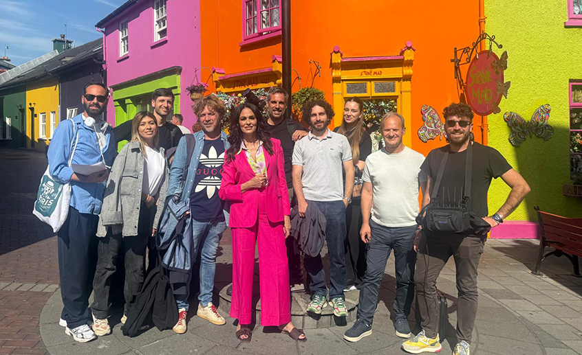 Group of people posing in front of brightly coloured shopfronts on a sunny street in a small Irish town.