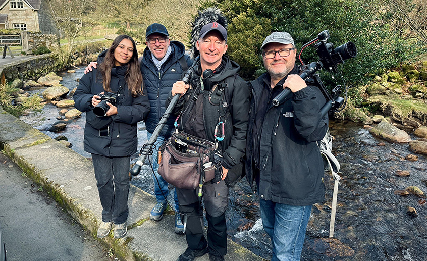 Four people standing together outdoors beside a rocky stream, holding camera and audio equipment.