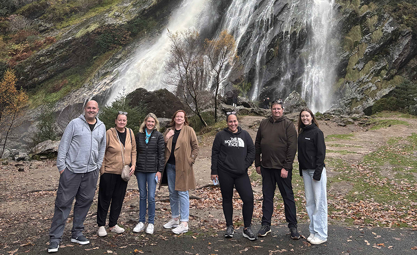 A group of six people standing outdoors in front of a waterfall