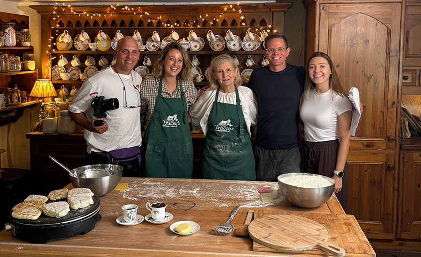 Five people standing behind a wooden kitchen counter with baking tools and dough, wearing aprons in a cozy, rustic kitchen lined with teacups and warm string lights.