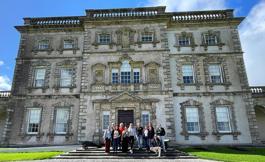 Group of people standing in front of a large historic stone building with ornate windows and architectural details under a blue sky.