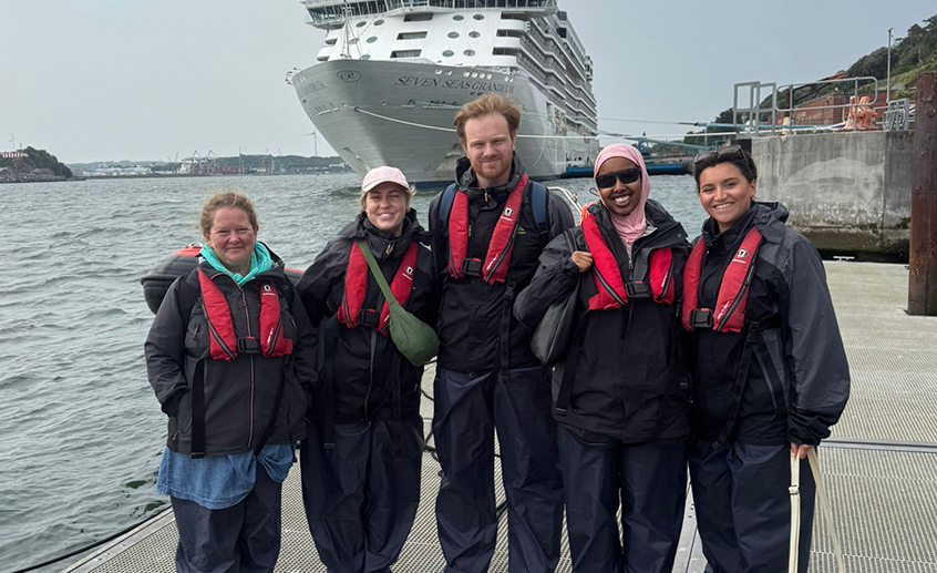 Five people standing in a harbour with a ship on the sea in the background.