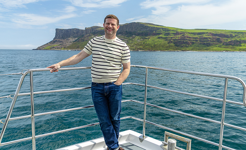 Person standing on a boat deck with calm sea and rugged green coastal cliffs in the background.