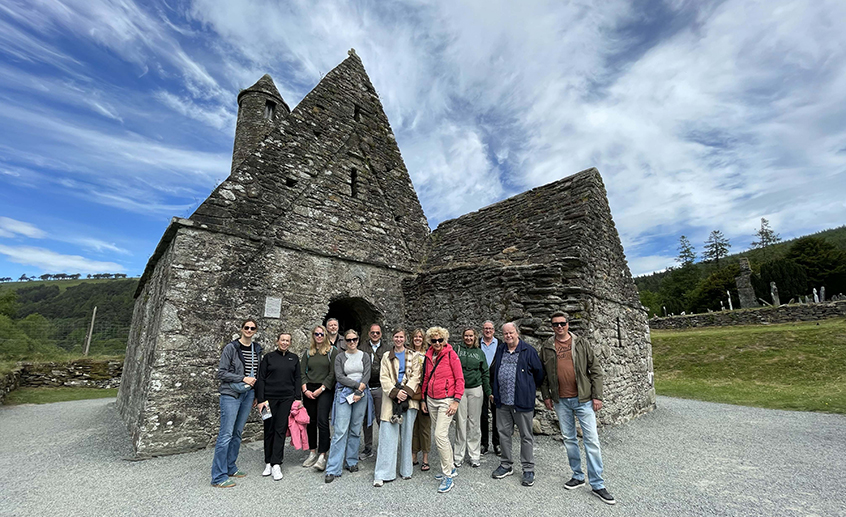 Group of visitors standing in front of an ancient stone church ruin under a blue sky with streaked clouds.