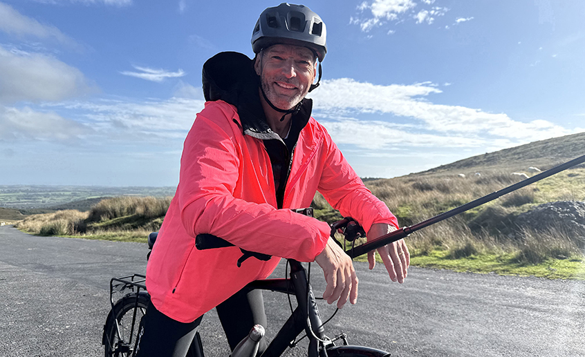 A cyclist in a bright pink jacket pauses on a rural road with rolling hills and a blue sky in the background.