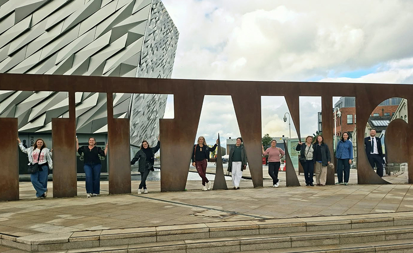 Group standing within the Titanic Belfast sign outside Titanic Belfast, with the museum building in the background.
