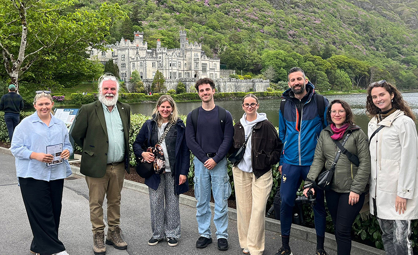 Group of eight people standing by a lakeside with a historic castle and lush green hillside in the background.