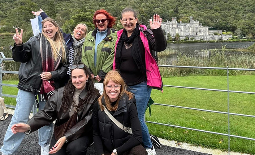 Six people pose beside a lakeside railing with Kylemore Abbey and wooded hills visible in the background.
