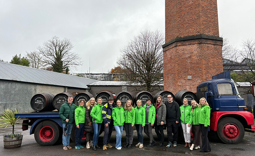 Group of people wearing green jackets standing outside beside a vintage truck loaded with barrels, in front of a tall brick chimney.