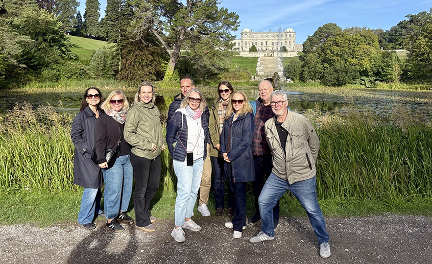 Group of travel industry delegates standing by a lakeside with a grand country house and landscaped grounds in the background.