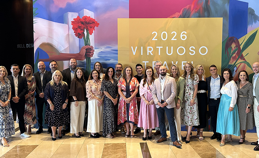 Group photo of tourism and travel industry representatives standing in front of a colourful mural and a sign reading “2026 Virtuoso Travel,” at an indoor event venue.