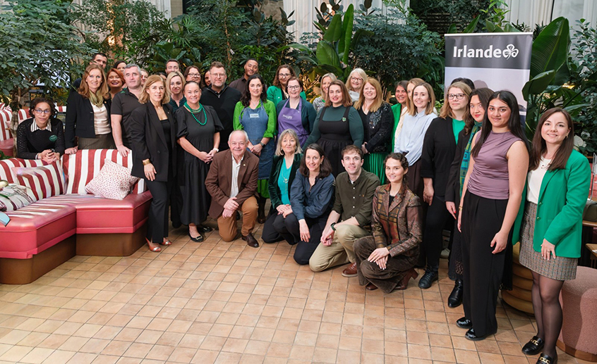 A large group of people gathered indoors for a formal event, standing and sitting together in front of lush greenery and an ‘Irlande’ banner.