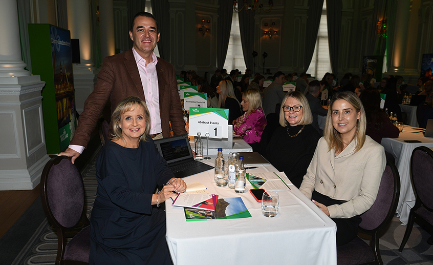 Group shot of four people sitting at a table with brochures, water bottles and a laptop; sign reads 