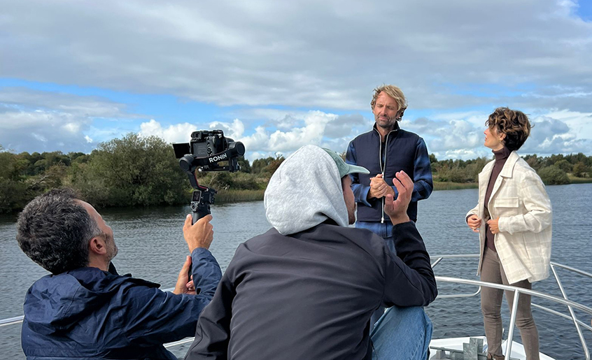 Four people standing filming on a boat on a river.