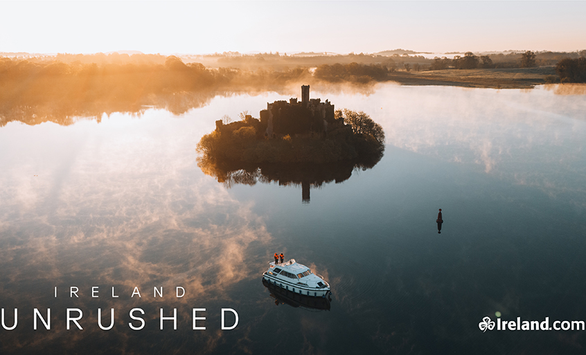 Aerial view of a small castle island reflected in calm water at sunrise, with a boat passing nearby