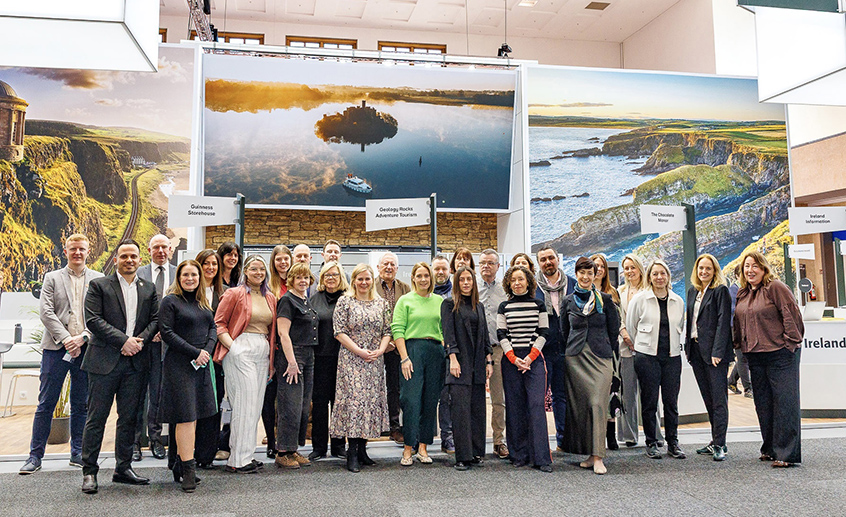 Group of people standing together at an indoor event, with scenic Ireland landscape displays in the background.