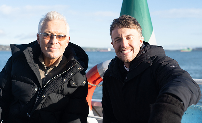 Two people in dark jackets on a boat, with the sea, an Irish flag and a lifebuoy visible in the background.