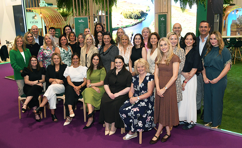Group of approximately 30 people posed for a team photo at a tourism exhibition stand, with green “Meet in Ireland” branding and scenic Irish imagery in the background.