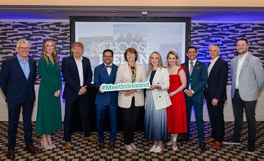 Group of ten people posing in front of a presentation screen, holding a “#MeetInIreland” sign at an event.
