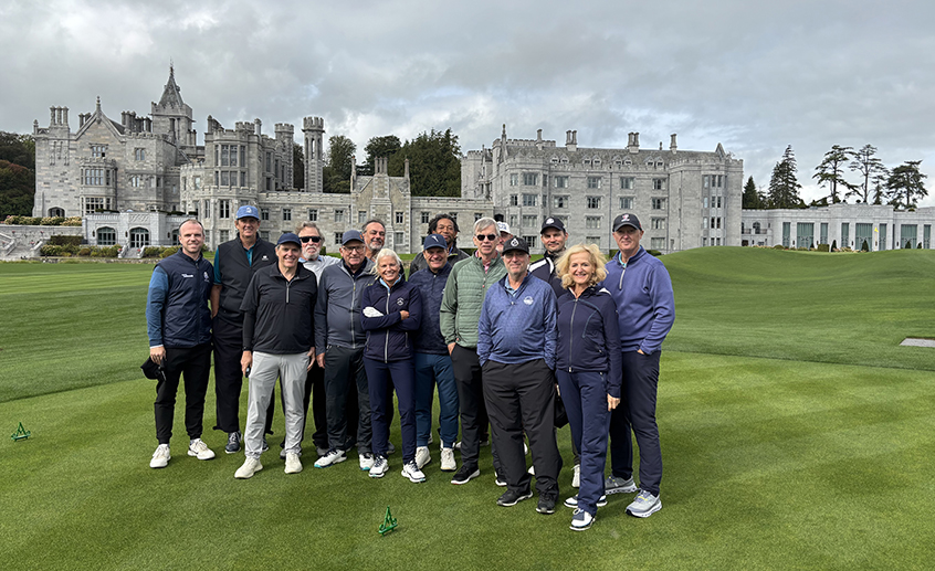 Group of golf journalists standing on a golf course in front of a large castle-style estate under cloudy skies.