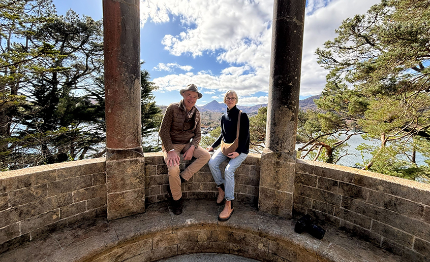 Two people sitting on a stone bench overlooking a scenic view of trees, water and mountains.