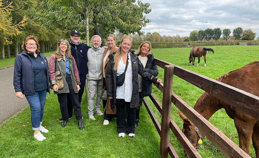 A group shot of seven people standing on grass beside a field with two horses grazing in it