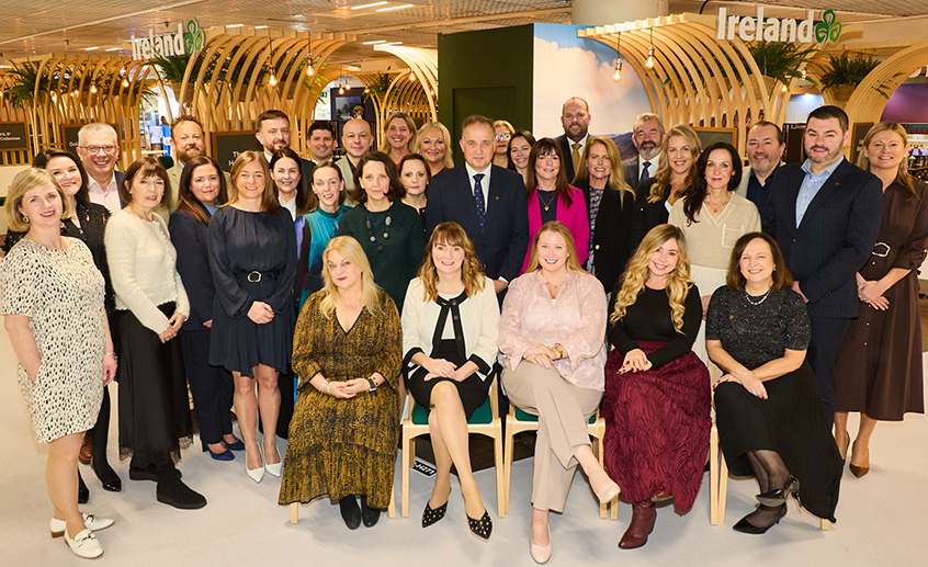 Group photo of people gathered at an indoor event space featuring a modern wooden arch design and Ireland branding in the background.