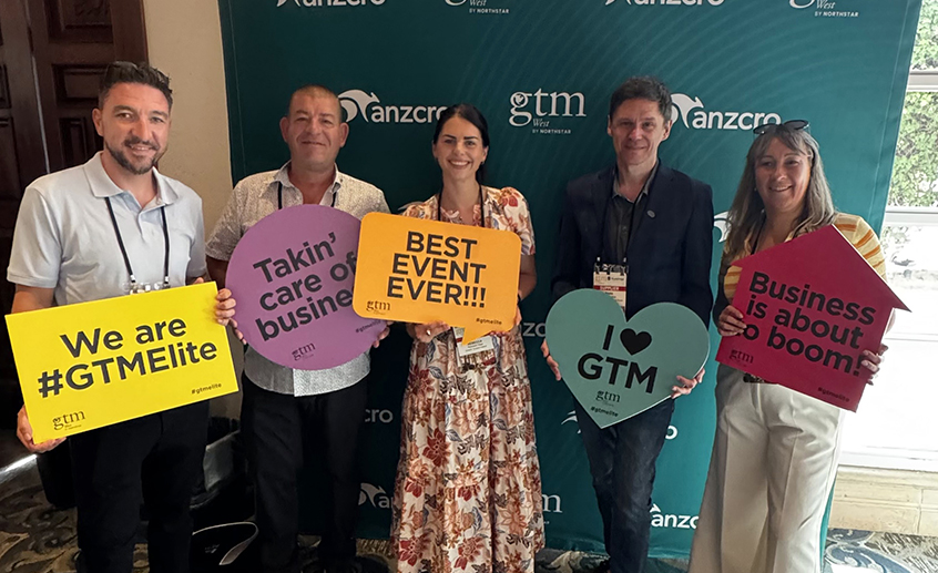 Five attendees pose in front of a GTM West branded backdrop, holding colourful promotional signs at a tourism industry event in California.