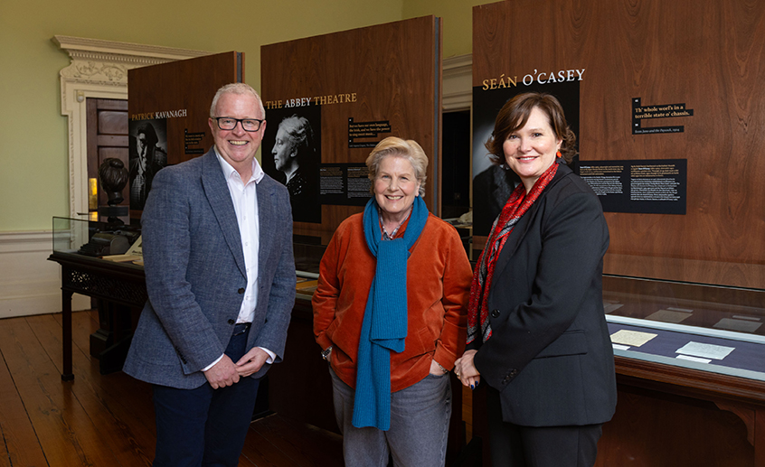 Three people standing in a museum-style exhibition space with display cases and Irish literary panels in the background.
