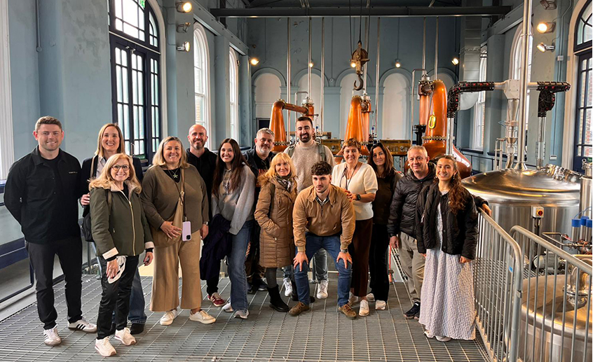 Group of visitors standing inside a distillery, with copper stills and industrial equipment in the background.