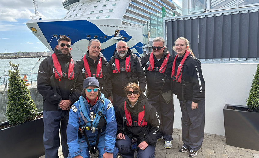Group of seven people in black waterproof gear and red life jackets posing by a harbor, with a large cruise ship docked in the background.