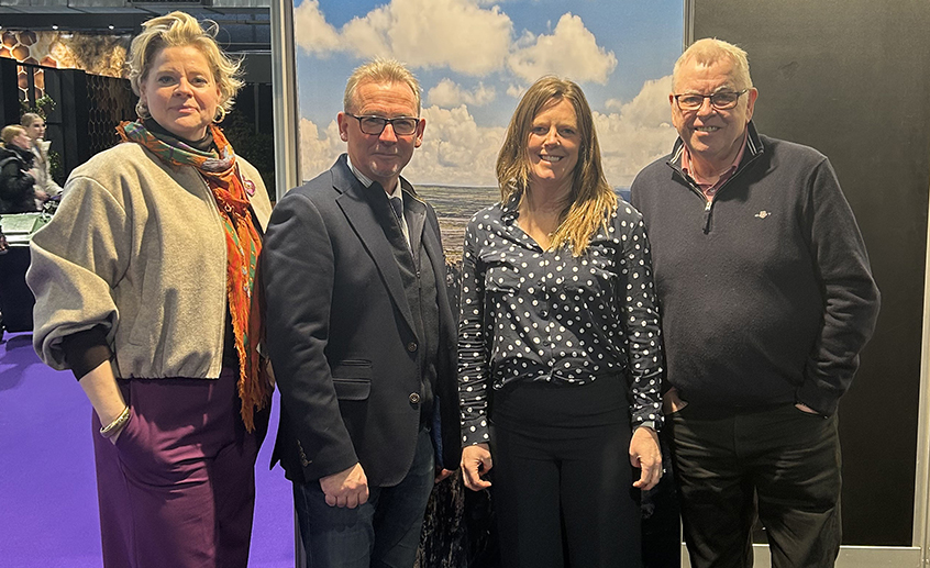 A group of four people standing together indoors in front of a scenic backdrop featuring a cloudy sky and shoreline.