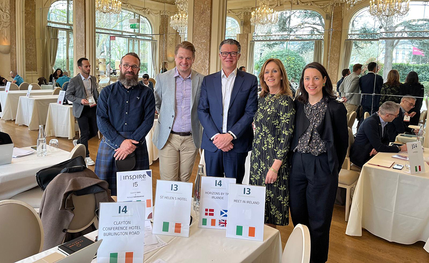 A group of people standing behind numbered meeting tables in an elegant, bright event space with large windows and chandeliers. Signs on the tables display venue names and small national flags.