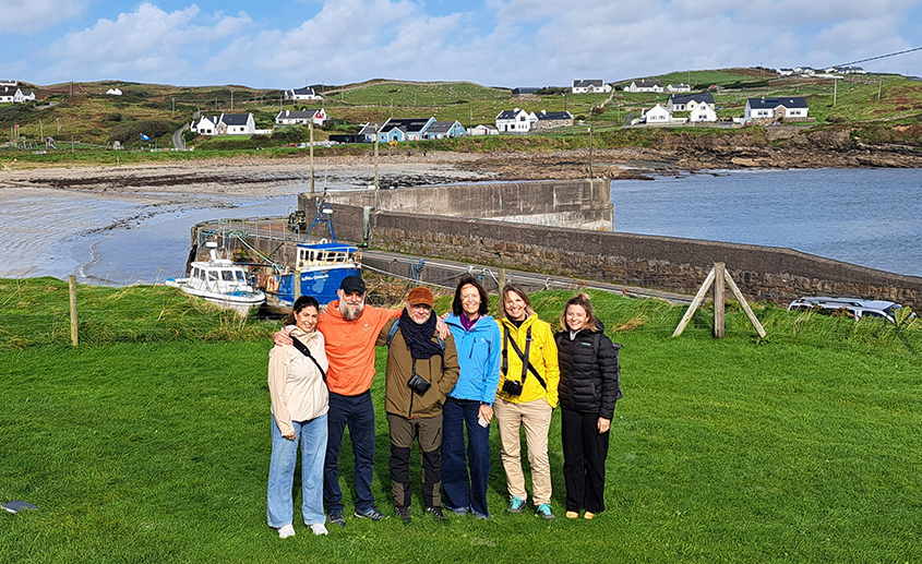 Group of travel journalists standing on a grassy hillside overlooking a small harbour and coastal village on Ireland’s rugged west coast.