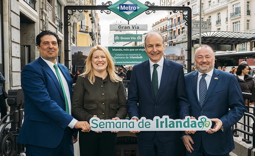 Four people holding a ‘Semana de Irlanda’ sign at the Gran Vía Metro entrance during an Ireland-themed event.