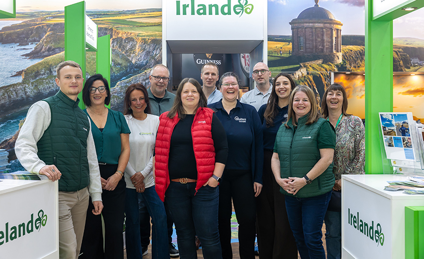 Group of people standing together at an Ireland-themed promotional booth featuring scenic imagery and tourism displays