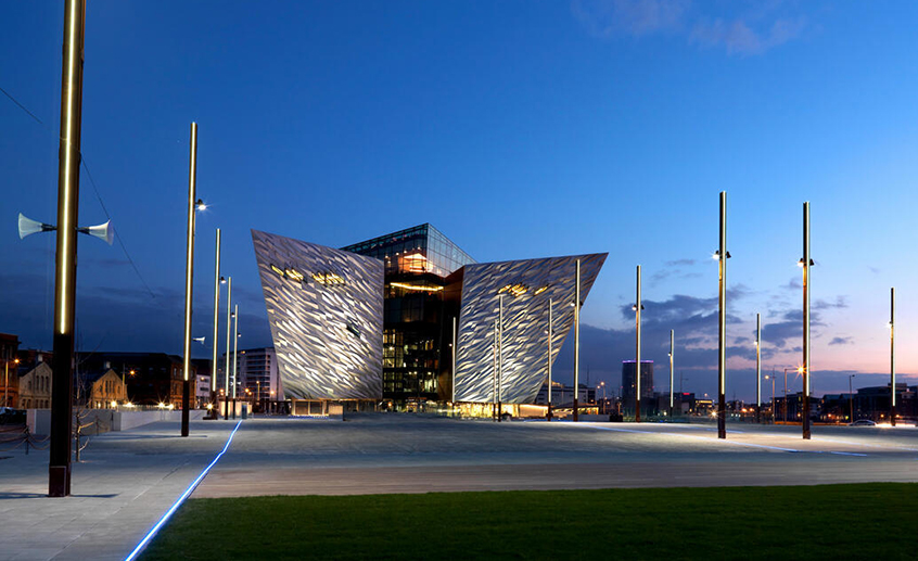 Titanic Belfast illuminated at dusk, with its striking angular façade and open plaza in the foreground.