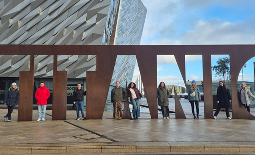 Group of people standing spaced out along a large ‘TITANIC’ metal sign outside the Titanic Belfast museum.