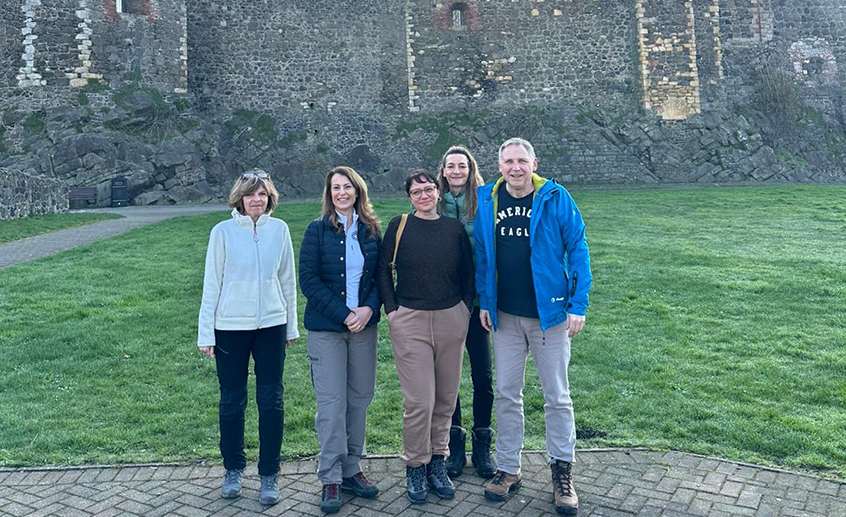 Five people standing outdoors in front of a stone wall and grassy area, posing for a group photo.