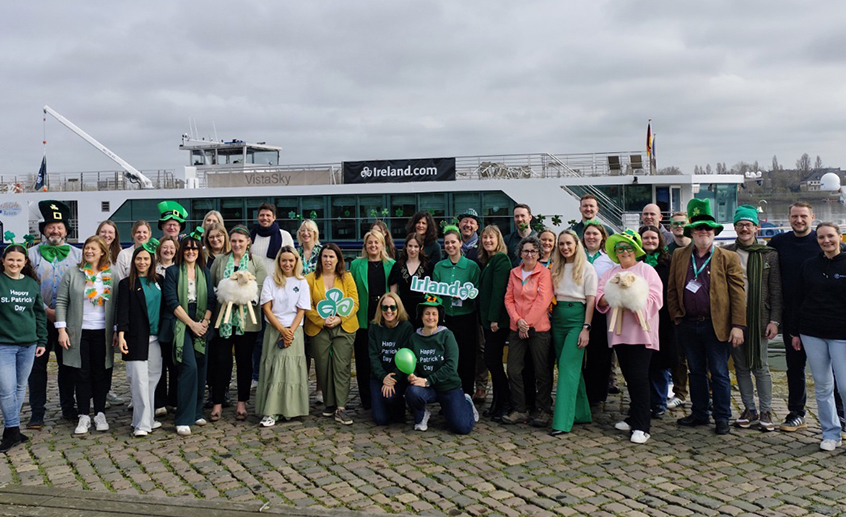 A large group of people dressed in green St. Patrick’s Day–themed clothing posing together on a cobblestone waterfront in front of an Ireland-branded boat.