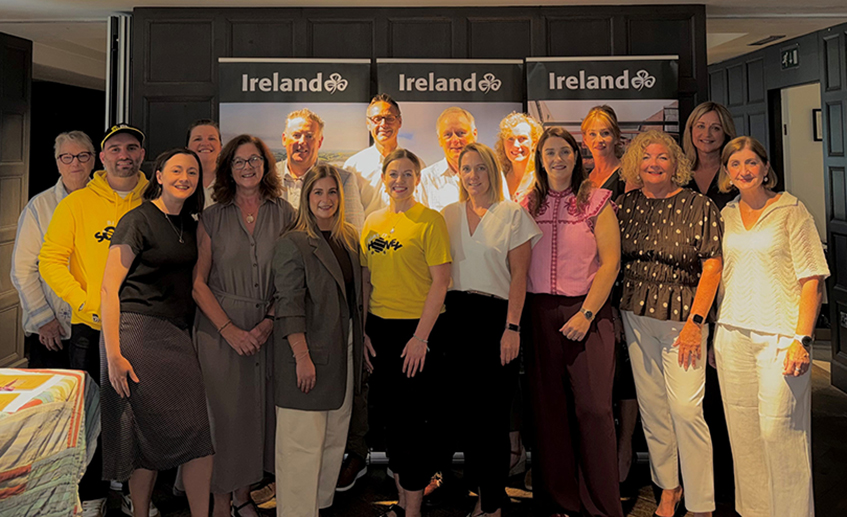 Group photo of event attendees standing in front of Ireland-branded display banners at an indoor Tourism Ireland gathering.