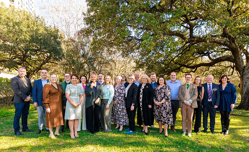 Group of people standing together outdoors on a grassy lawn, with large trees in the background on a sunny day