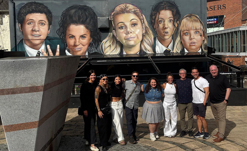 Group of people posing in front of a large mural featuring five painted faces on a city building in Northern Ireland.
