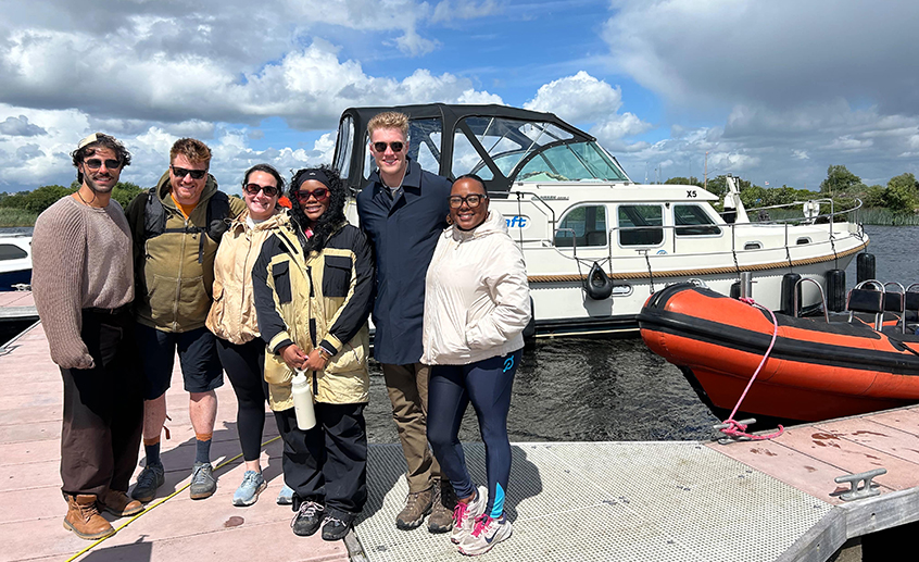 Six people standing on a dock beside a boat on a lake under a partly cloudy sky.