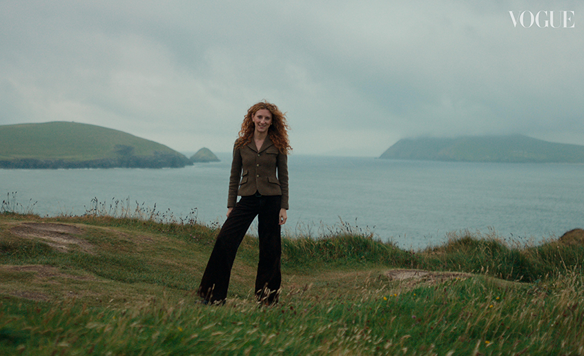 Person standing on a grassy coastal cliff overlooking the sea and distant islands under a cloudy sky.