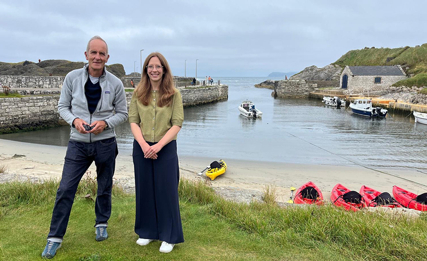 Two people posing in front of a harbor with kayaks lined along the shore.