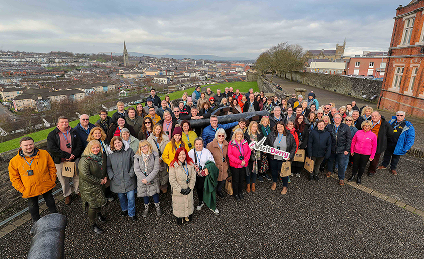 Large group of people gathered outdoors on a walkway overlooking the city of Derry-Londonderry, with buildings and hills visible in the background.
