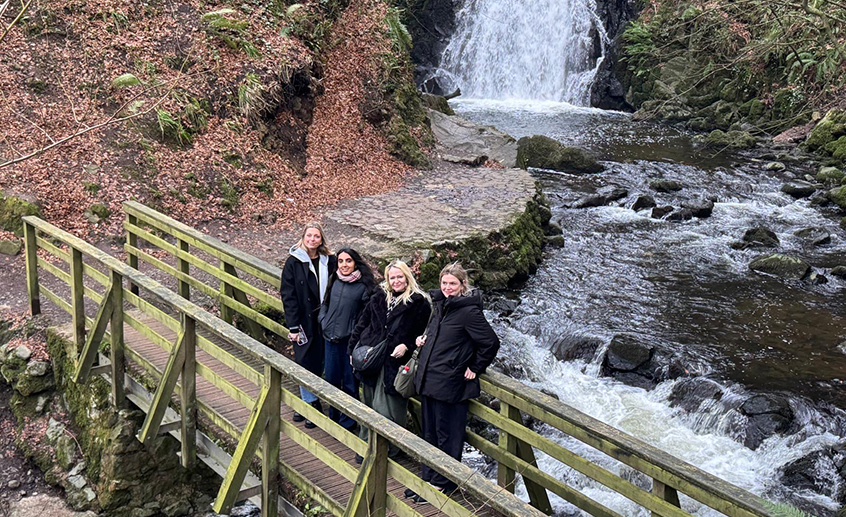 Four people standing on a wooden bridge in front of a flowing river and waterfall in a forest setting